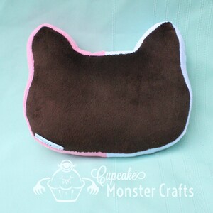 Puede incluir: Una almohada marr&oacute;n con forma de gato con ribetes rosas y blancos. La almohada est&aacute; hecha de un material suave y afelpado y presenta un dise&ntilde;o sencillo. El texto "Cupcake Monster Crafts" es visible en la parte inferior.
