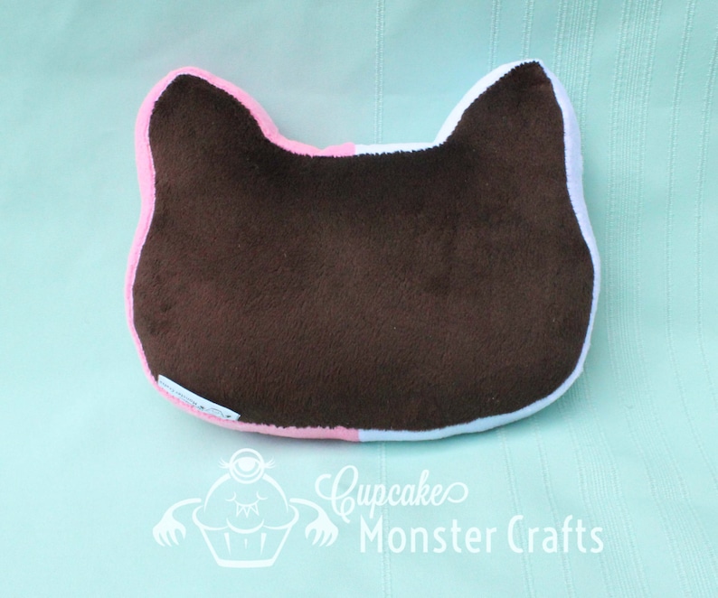 Puede incluir: Una almohada marr&oacute;n con forma de gato con ribetes rosas y blancos. La almohada est&aacute; hecha de un material suave y afelpado y presenta un dise&ntilde;o sencillo. El texto "Cupcake Monster Crafts" es visible en la parte inferior.