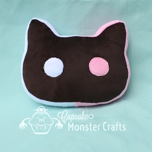 Puede incluir: Un coj&iacute;n con forma de gato, marr&oacute;n con detalles blancos y rosas para los ojos y las orejas. Tiene un borde azul claro y est&aacute; sobre un fondo azul claro. El texto "Cupcake Monster Crafts" es visible.