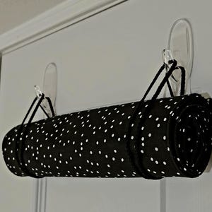 May include: A black fabric roll featuring a white polka dot pattern, suspended horizontally by two black cords from two clear adhesive wall hooks. The cylindrical textile item is tightly rolled and secured with black string loops.