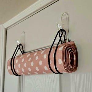 May include: A rolled-up pink fabric with white polka dots is suspended from a white door using black cords and clear adhesive hooks. The fabric is neatly rolled, showcasing the pattern and texture.