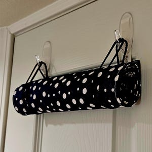 May include: A cylindrical fabric roll featuring a black background with a white polka dot pattern. The roll is suspended horizontally by two black braided cords, each looped over a clear adhesive plastic hook attached to a flat vertical surface.