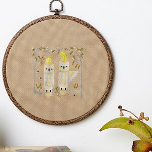 May include: A beige embroidery hoop with a cross-stitch design of two yellow cockatoos on a branch. The hoop is hanging on a white wall. Two books are on a table with a green and white patterned tablecloth. The books are titled "Koala" and "The Secret Life of Wombats".