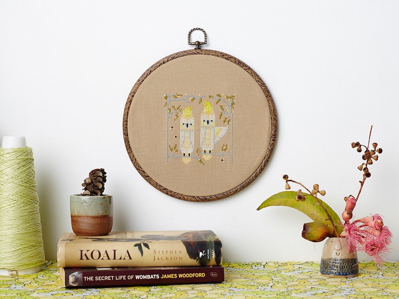 May include: A beige embroidery hoop with a cross-stitch design of two yellow cockatoos on a branch. The hoop is hanging on a white wall. Two books are on a table with a green and white patterned tablecloth. The books are titled "Koala" and "The Secret Life of Wombats".