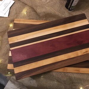 May include: A wooden cutting board with a striped design. The board is made of different types of wood, including walnut, maple, and purpleheart. The board is approximately 18 inches long and 12 inches wide.