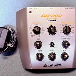 May include: A silver Zoom GM-200 Guitar Amp Modeler with black control knobs, labeled settings for amp selection, effects, and output correction, and a black power adapter with a cord. The device features gain, ZNR, master level, treble, middle, and bass controls.