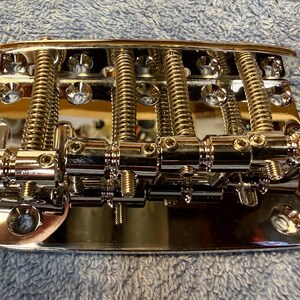 May include: A shiny chrome bass guitar bridge with adjustable saddles and string height screws. The bridge has a rectangular base with multiple mounting holes and is designed to hold the strings in place.
