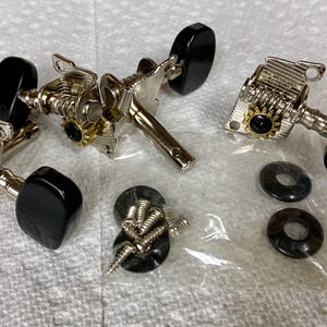 May include: A collection of guitar tuning machines with black tuning pegs, silver metal components, and gold-colored gears. Also included are screws, washers, and a small plastic bag.