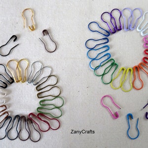 May include: A collection of colourful safety pins arranged in a circle. The pins are in various colours, including red, orange, yellow, green, blue, purple and white. There are also some gold and silver safety pins.