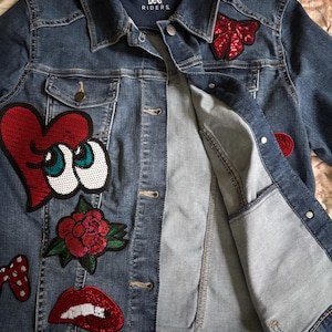 May include: A denim jacket with various sequined patches, including a heart with eyes, a rose, lips, and a bow. The jacket is a medium wash blue with the "Lee Riders" logo visible on the collar.