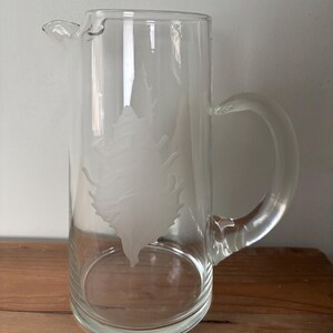 May include: A clear glass pitcher with a handle and a spout. The pitcher has a frosted design of a tree on the front. The pitcher is designed for serving beverages.