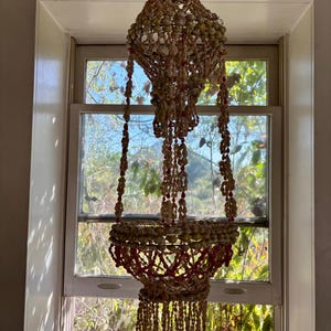 May include: A hanging seashell chandelier with a tiered design. The chandelier features a top sphere, a central basket, and dangling strands, all adorned with various seashells and red accents. It hangs in front of a window with a view of greenery.