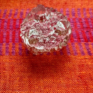 May include: A clear, textured glass votive candle holder. The candle holder is round with a rough, uneven surface. The background is a vibrant orange and red woven fabric with purple stripes.