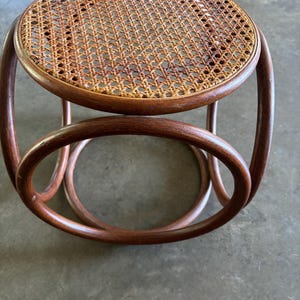 May include: A round stool featuring a circular seat with a woven cane pattern in a light tan color. The frame is constructed from dark brown bentwood, consisting of a circular top rim supported by two large, interlocking circular base structures.