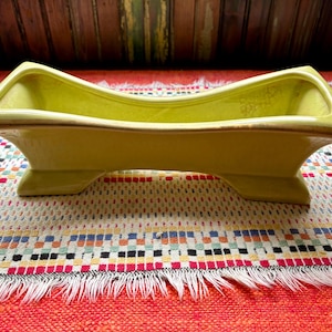 May include: A chartreuse-colored ceramic planter with a curved design. The planter has a rectangular shape with a slightly curved top and is supported by four feet. The rim has a brown trim. The planter is sitting on a colorful patterned textile.