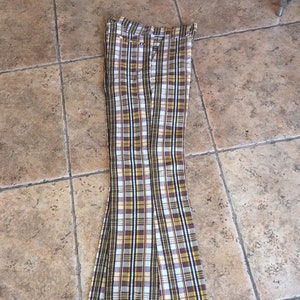 May include: A pair of high-waisted trousers featuring a prominent plaid pattern in shades of mustard yellow, brown, white, and black. The pants have a flared leg silhouette, a structured waistband with belt loops, and a front zipper closure.