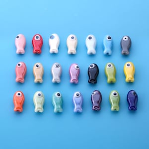 May include: A collection of twenty-one small, smooth, fish-shaped beads arranged in three rows. Each bead features a glossy finish in various solid colors, including red, blue, yellow, green, pink, black, and white, with a single eye detail.