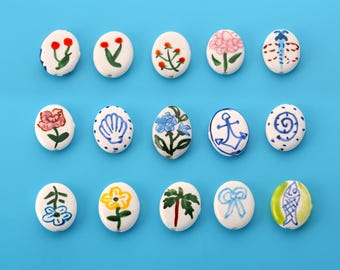 May include: A collection of fifteen oval-shaped white ceramic beads arranged in three rows. Each bead features a unique hand-painted design, including various flowers, a seashell, an anchor, a dragonfly, a bow, and a fish in blue, green, red, and yellow.