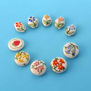 Könnte beinhalten: Eine Sammlung von zehn weißen Keramikperlen, die in einem Kreis angeordnet sind. Jede Perle zeigt handbemalte Motive, darunter verschiedene bunte Blumen in Rot, Blau, Gelb und Rosa mit grünen Stielen und Blättern sowie eine rote Erdbeere.