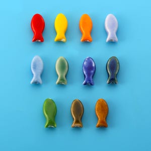 Könnte beinhalten: Eine Sammlung von elf kleinen, glatten, fischförmigen Perlen, die in drei Reihen angeordnet sind. Die Perlen haben eine glänzende Oberfläche und sind in verschiedenen Unifarben erhältlich, darunter Rot, Gelb, Orange, Helllila, Hellblau, Salbeigrün, Dunkelblau, Blaugrün, Limettengrün, Braun und Hellbraun.