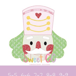 May include: A digital embroidery design of a nutcracker face. The design features a white face with a white mustache, red cheeks, and a pink and white checkered hat with a red heart. The text "Sweet CC EMBROIDERY DESIGNS" is at the bottom.