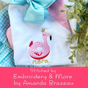 Puede incluir: Top blanco y rosa con un diseño de flamenco. El flamenco es rosa con una corona dorada y el nombre "Nora" en letras doradas. Un gran lazo azul está en la parte superior. El texto "Stitched by Embroidery & More by Amanda Brazeau" está en la parte inferior.