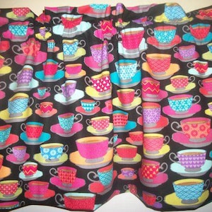 May include: A black fabric with a repeating pattern of colorful teacups and saucers. The teacups are in various colors, including pink, blue, red, and purple, with yellow saucers. The fabric is suitable for home decor.