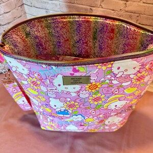 Pode incluir: Uma sacola de tecido rosa com fecho de z&iacute;per, com um padr&atilde;o repetido de personagens de desenhos animados e flores. O forro interno tem um design abstrato colorido. Uma al&ccedil;a de pulso &eacute; presa na lateral.