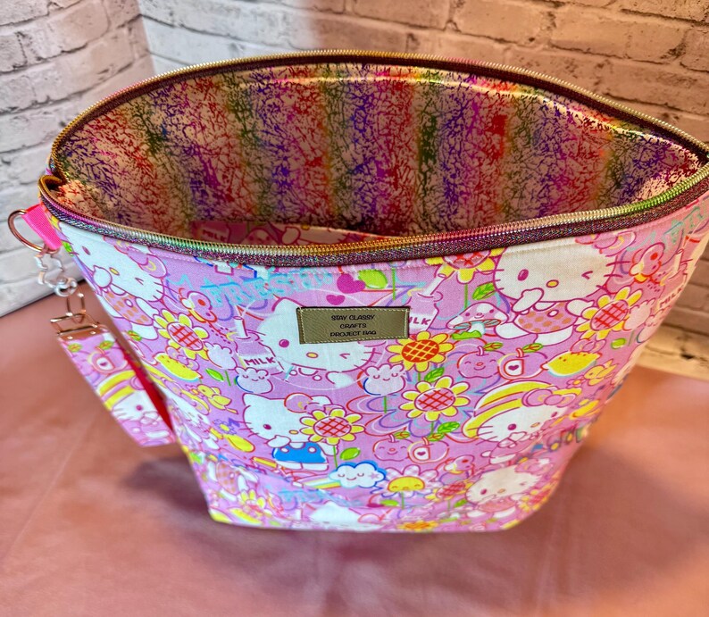 Pode incluir: Uma sacola de tecido rosa com fecho de z&iacute;per, com um padr&atilde;o repetido de personagens de desenhos animados e flores. O forro interno tem um design abstrato colorido. Uma al&ccedil;a de pulso &eacute; presa na lateral.