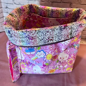 Pode incluir: Uma sacola de tecido com um padr&atilde;o Hello Kitty rosa e branco. A sacola tem uma borda estampada abstrata e colorida e um interior rosa. A sacola est&aacute; aberta, revelando o bolso interno.