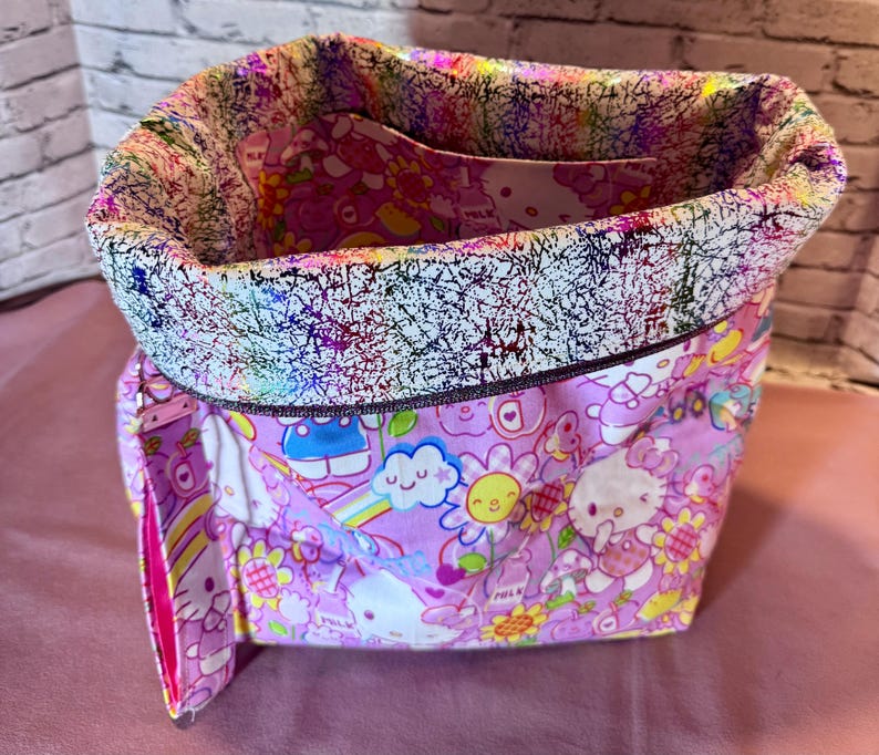 Pode incluir: Uma sacola de tecido com um padr&atilde;o Hello Kitty rosa e branco. A sacola tem uma borda estampada abstrata e colorida e um interior rosa. A sacola est&aacute; aberta, revelando o bolso interno.
