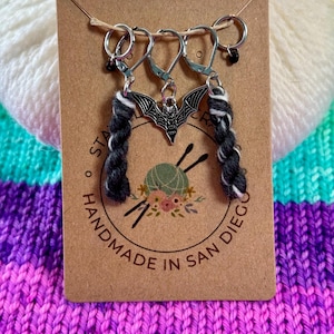 May include: A set of three silver stitch markers with black and white yarn tails. The markers feature a bat charm and the text "Handmade in San Diego".