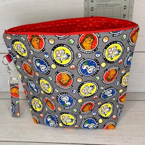 Puede incluir: Una bolsa de cremallera gris con forro rojo. La bolsa está decorada con un patrón repetido de círculos coloridos con personajes de Star Wars, incluyendo Chewbacca y D-O. El texto "Rolling Behind the Resistance Crew" está impreso en los círculos.