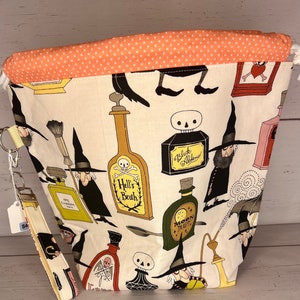 May include: A white fabric drawstring bag with a peach polka dot lining. The bag features a black and white print of witches, potions, and skulls. The text on the potions includes "Hell's Broth", "Black Alchemy", and "Moon Mist".