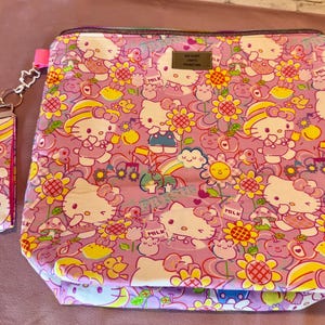 Pode incluir: Uma bolsa rosa com z&iacute;per e uma al&ccedil;a de pulso combinando, ambas cobertas por um padr&atilde;o repetido de personagens Hello Kitty, flores, arco-&iacute;ris e outros elementos l&uacute;dicos. A bolsa tem uma pequena etiqueta de metal.