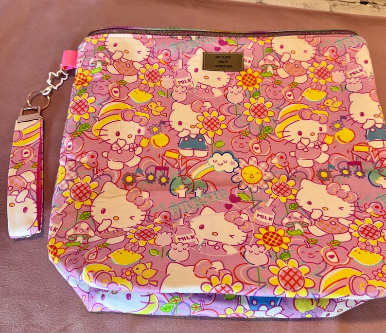 Pode incluir: Uma bolsa rosa com z&iacute;per e uma al&ccedil;a de pulso combinando, ambas cobertas por um padr&atilde;o repetido de personagens Hello Kitty, flores, arco-&iacute;ris e outros elementos l&uacute;dicos. A bolsa tem uma pequena etiqueta de metal.