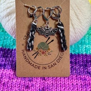 May include: Three black and white yarn stitch markers with a silver bat charm. The stitch markers are on a brown card with the text "Handmade in San Diego".