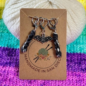 Op de afbeelding: Een set van drie zilveren stekenmarkeerders met zwart-witte garenaccenten. Eén markeerder heeft een vleermuisbedel. De kaart leest "HANDMADE IN SAN DIEGO" met een garen- en naaldlogo. Een bol witte wol staat achter de kaart.
