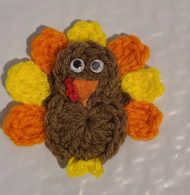 May include: A handmade turkey decoration made of brown yarn with googly eyes, an orange beak, and a red wattle. The turkey has yellow and orange yarn feathers. A festive, handcrafted item for Thanksgiving or fall decor.