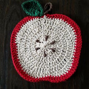 May include: A handmade crochet apple-shaped trivet. The trivet features a red border, a white and beige interior, and a green leaf with a brown stem. The design mimics a cross-section of an apple, perfect for kitchen decor.
