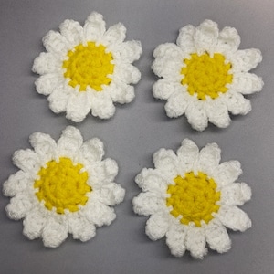 May include: Four crocheted white daisies with yellow centers. The daisies are arranged in a square pattern on a gray background.