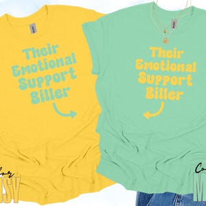 May include: Two t-shirts, one yellow and one mint green, are displayed. Each shirt features the text "Their Emotional Support Biller" in a contrasting color, with an arrow pointing to the right. The word "Color" and the color name are at the bottom.