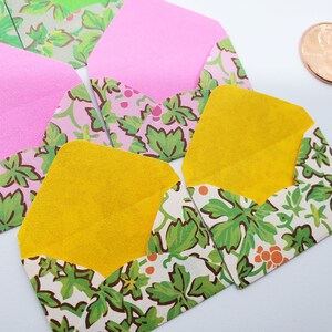 Könnte beinhalten: Eine Sammlung kleiner, farbenfroher Umschläge. Einige sind rosa oder gelb, mit gemusterten Klappen mit grünen Blättern und Beeren auf cremefarbenem Hintergrund. Eine Münze ist in der oberen rechten Ecke sichtbar.