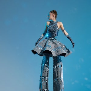 Puede incluir: Una mujer con un vestido de lentejuelas plateadas y pantalones a juego está de pie sobre zancos. Los zancos también están cubiertos de lentejuelas plateadas. Ella está posando contra un fondo azul.