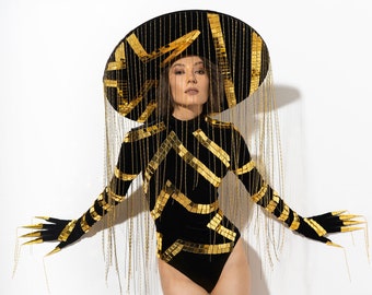 May include: A woman wearing a black bodysuit with gold sequins and a large black hat with gold fringe. The hat is decorated with gold sequins and has a wide brim.