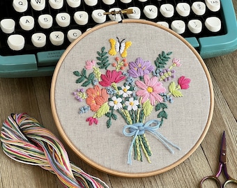 Puede incluir: Ramo floral bordado en un aro de madera. El colorido ramo presenta flores rosas, naranjas, moradas y blancas con una mariposa amarilla. Una cinta azul claro ata los tallos. Hilo de bordar y tijeras también están en la imagen.