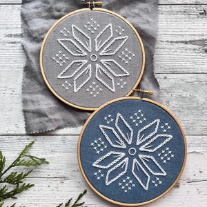 May include: Two embroidery hoops with white snowflake designs. The hoop on the left is gray and the hoop on the right is blue.