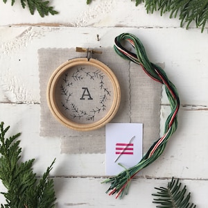 May include: A white embroidery hoop with a black embroidered design of a wreath of pine branches with the letter A in the center. The hoop is on a beige linen fabric with a skein of green, red, and white embroidery floss and a needle on a white card.