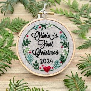 May include: A circular wooden embroidery hoop containing light gray fabric with hand-stitched black text reading Olivia's First Christmas 2024. The design features a green botanical wreath with small red berries and a red heart.