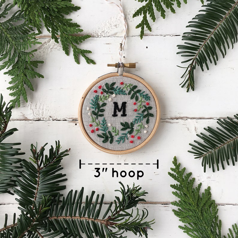 May include: A 3-inch embroidery hoop with a white linen fabric background. The fabric is embroidered with a green wreath with red berries and a white pearl border. The letter "M" is embroidered in the center of the wreath.